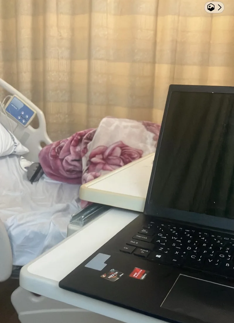 A laptop on a hospital bed table beside a bed during a period of illness in August 2025